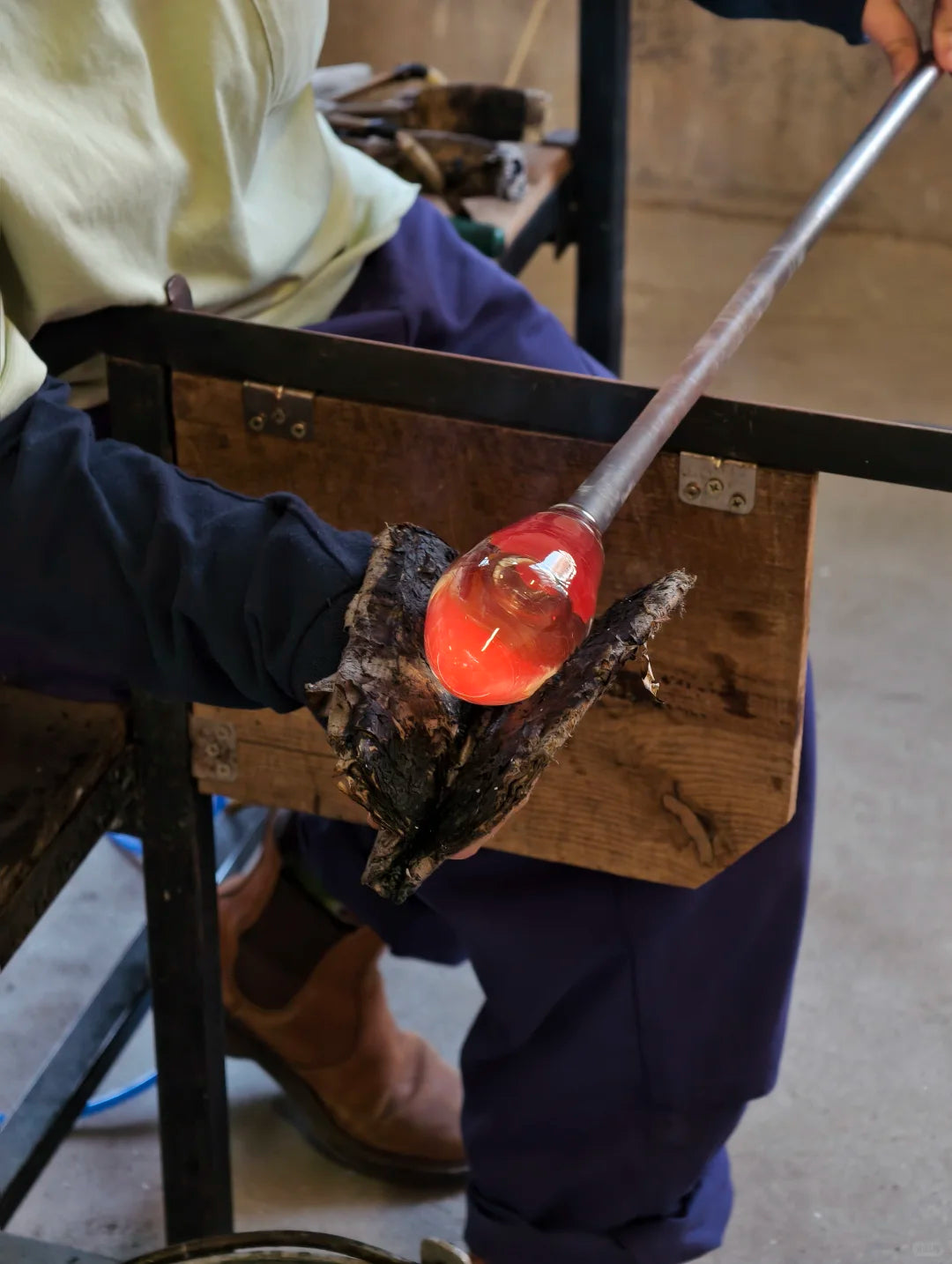A close-up of a glassblower's hands holding a glowing orange-red molten bubble on a metal blowpipe, mounted on a wooden stand in a workshop setting.